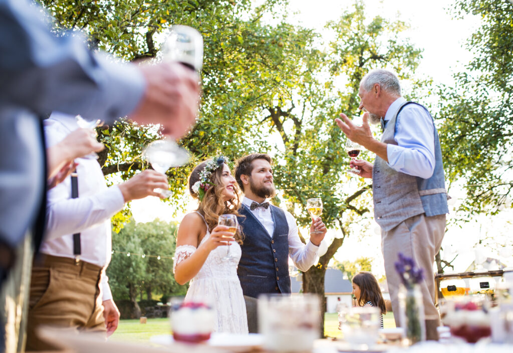 A senior man giving a wedding speech at a reception outside in the background.