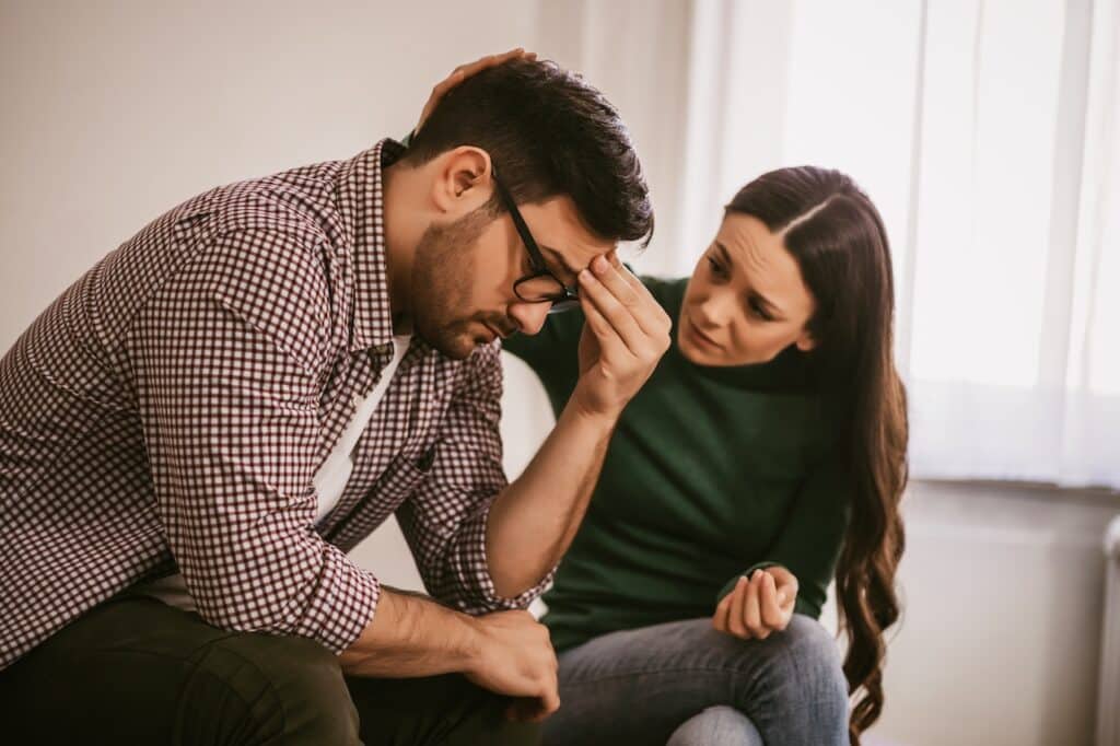 stressed man sitting at home with concerned looking wife who is trying to console him